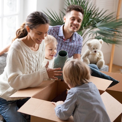 Happy family with two kids unpacking boxes after relocation moving into or settling in new home concept, excited small children helping parents with belongings sitting on sofa in living room together